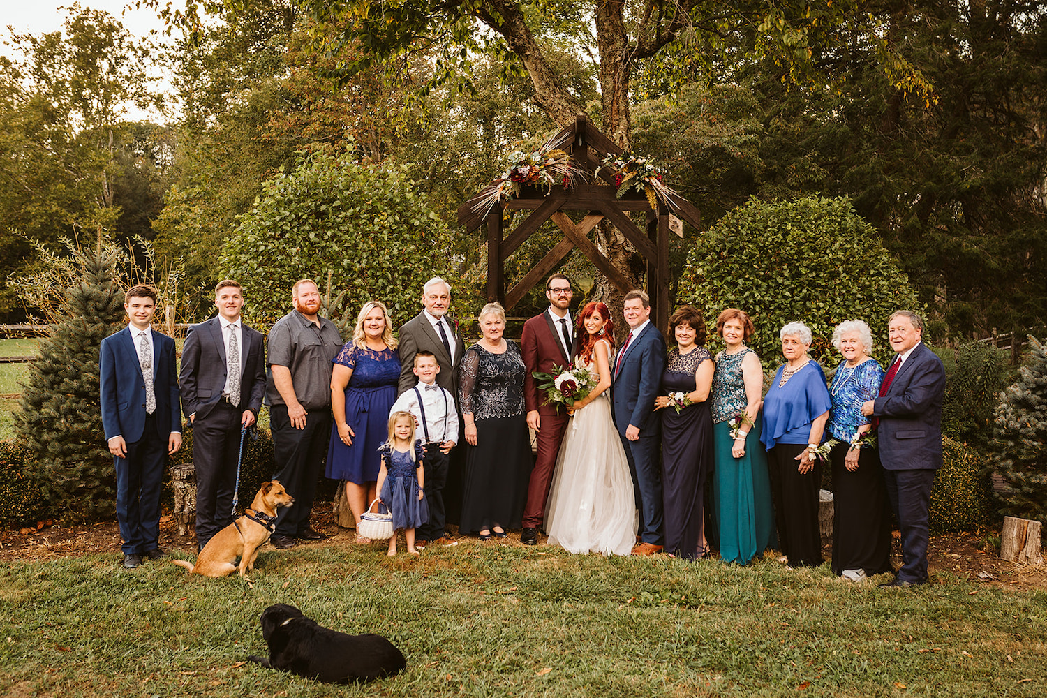 bride and groom pose with their family members and dogs in front of the wooden arch ceremony structure