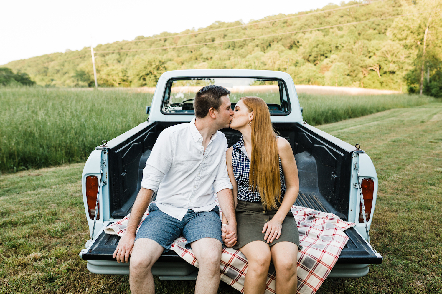 man and woman hold hands and kiss while sitting on bed of Ford truck in field of grass near Chattanooga
