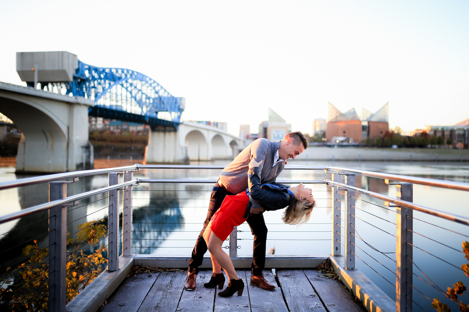 man in gray sweater dips woman in short red dress and black leather jacket on a pier near Chattanooga Market Street Bridge