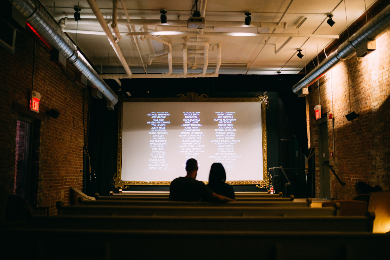 man and woman sit in church pew in a small theater with credits on the screen