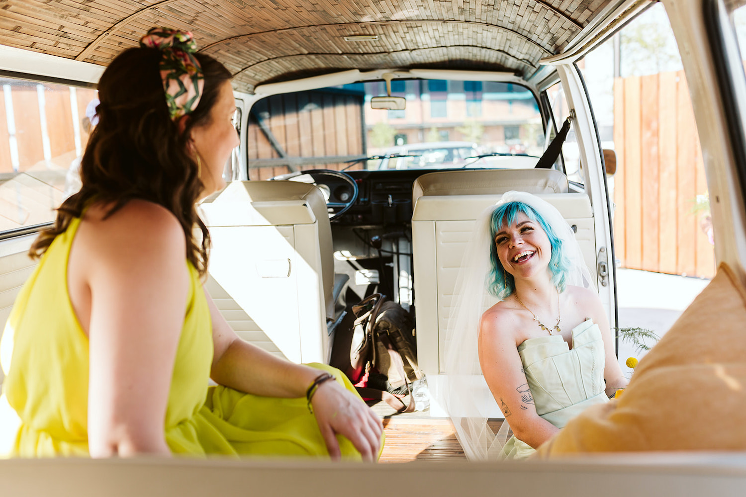 bride with blue hair and strapless dress laughs with woman in yellow dress inside a vintage Volkswagon bus