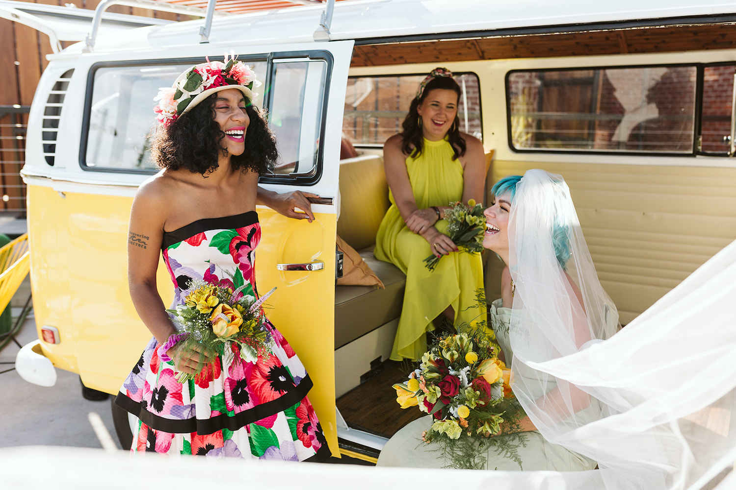 bride with blue hair laughs with woman in yellow dress and woman in bright floral dress and hat