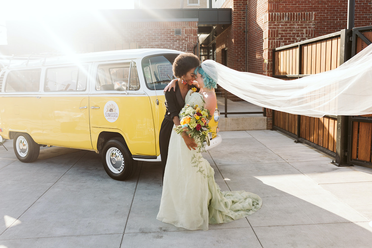 bride in black pantsuit hugs bride in strapless white gown at LGBTQ wedding in Chattanooga in front of yellow Volkswagon bus