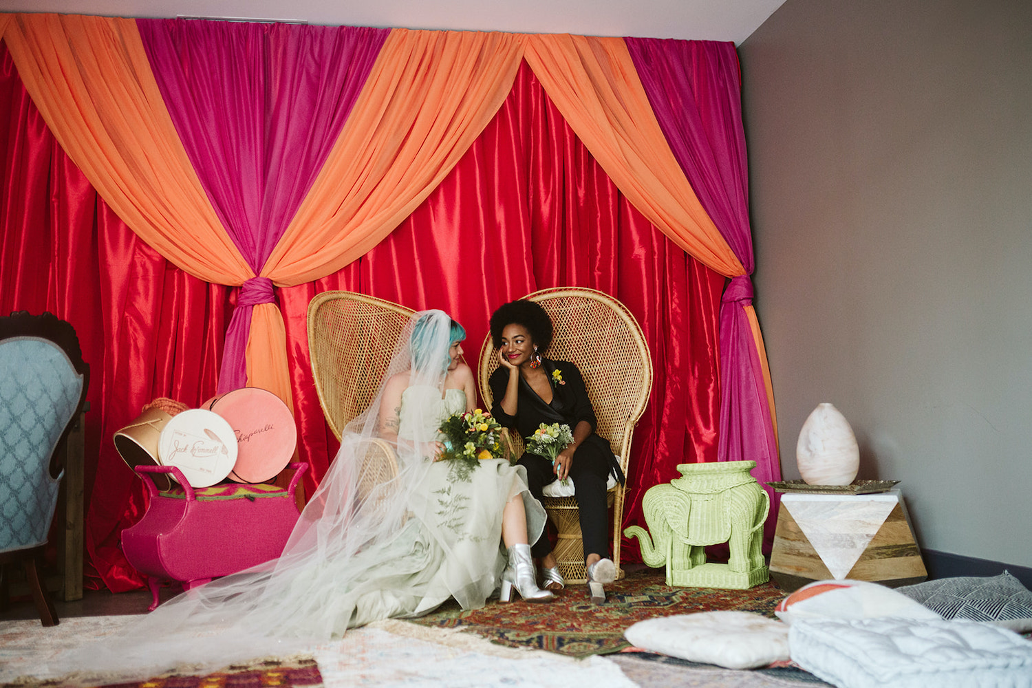 lesbian couple sits on peacock rattan chairs in front of bright wall draperies at Moxy Hotel at LGBTQ Wedding in Chattanooga