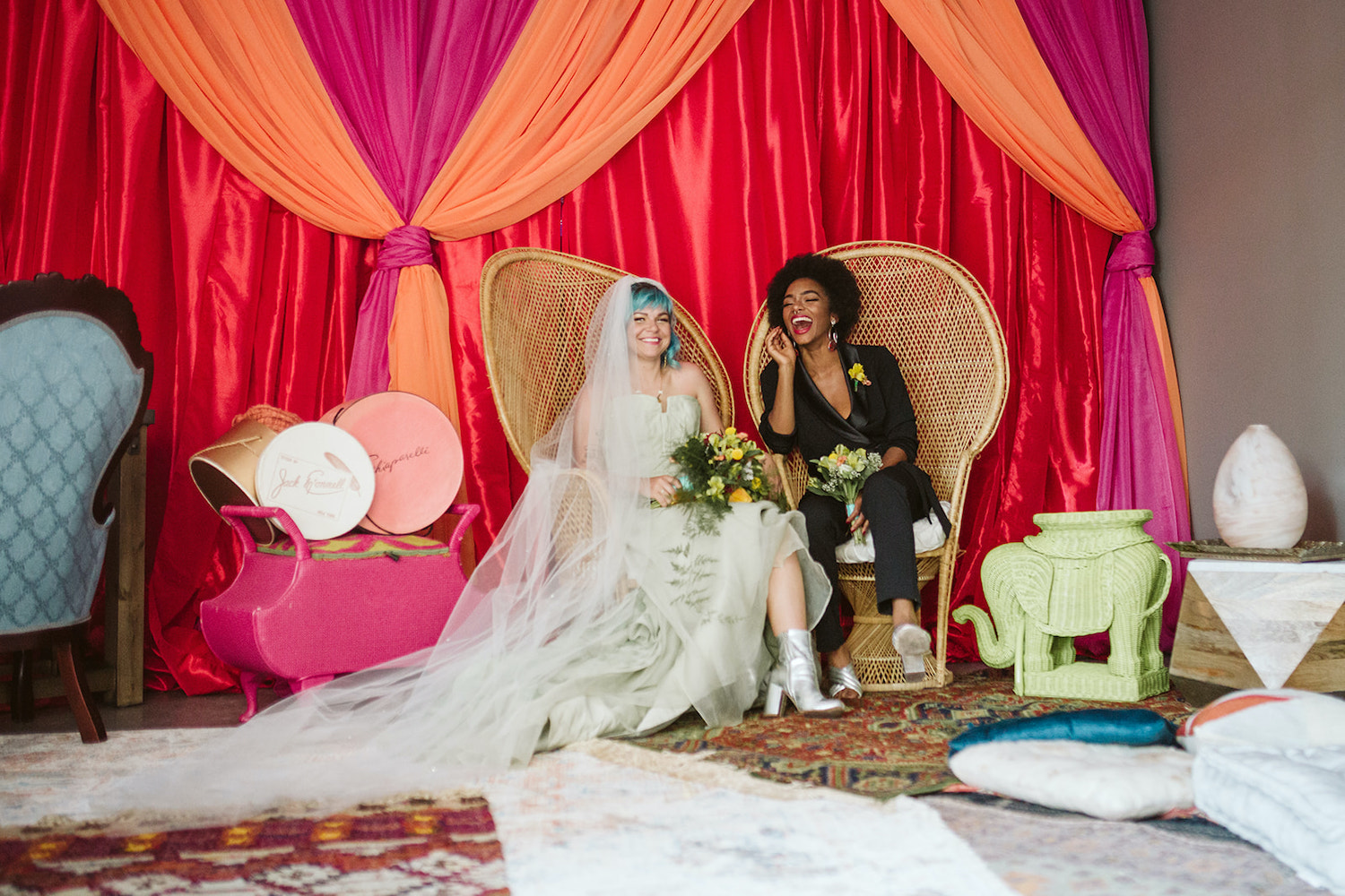 lesbian couple sits on peacock rattan chairs in front of bright wall draperies at Moxy Hotel at LGBTQ Wedding in Chattanooga