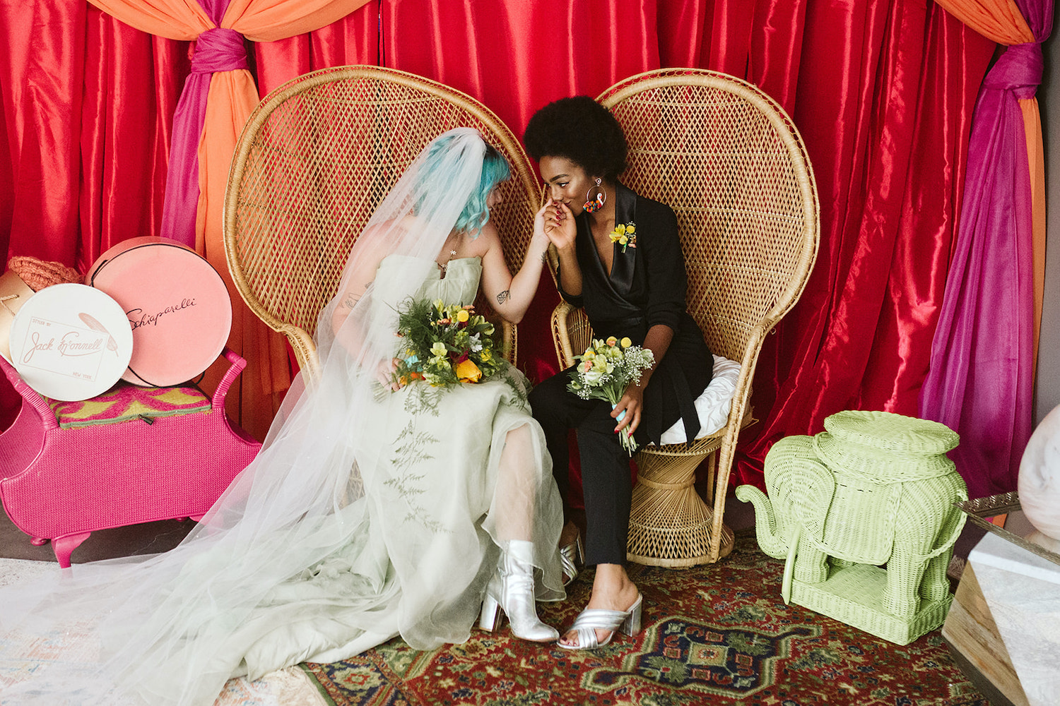 lesbian couple sits on peacock rattan chairs in front of bright wall draperies at Moxy Hotel at LGBTQ Wedding in Chattanooga