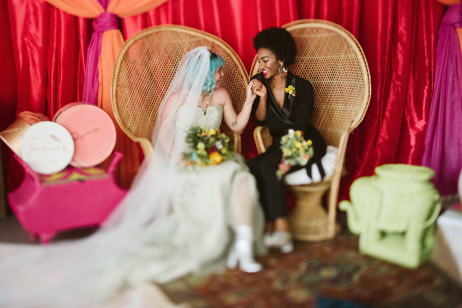 lesbian couple sits on peacock rattan chairs in front of bright wall draperies at Moxy Hotel at LGBTQ Wedding in Chattanooga