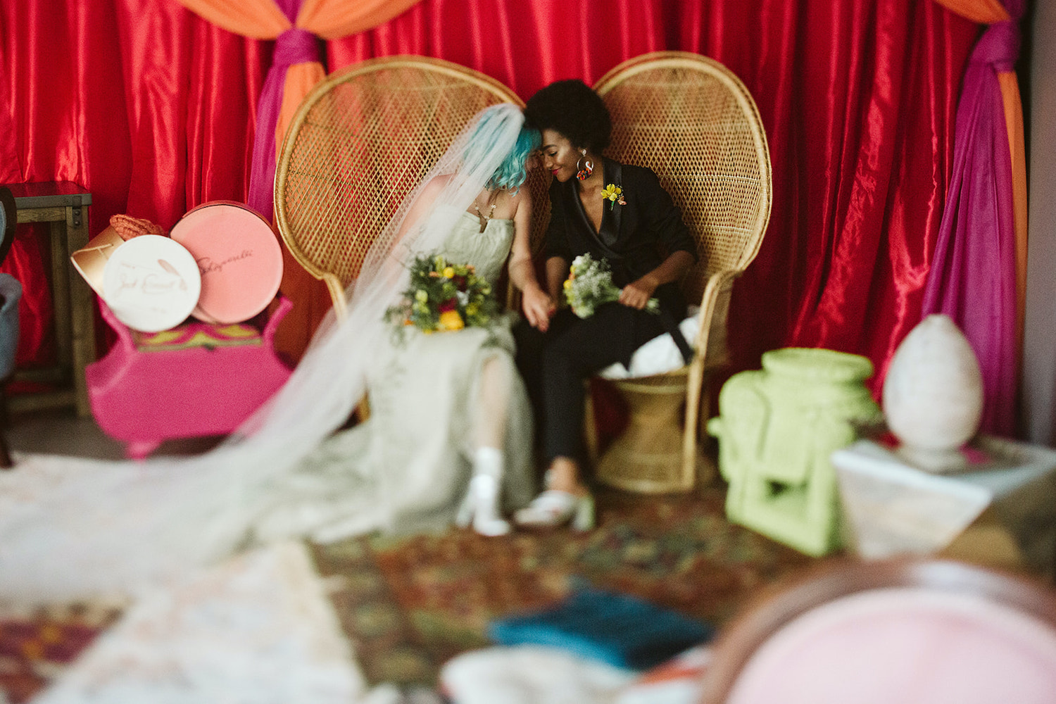 lesbian couple sits on peacock rattan chairs in front of bright wall draperies at Moxy Hotel at LGBTQ Wedding in Chattanooga