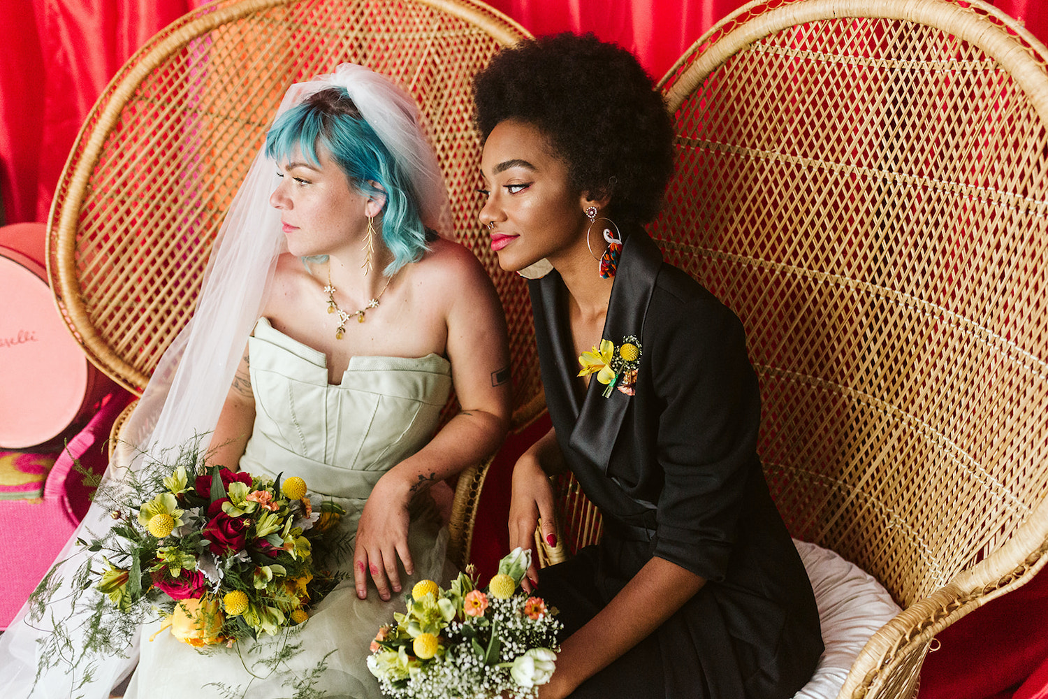 lesbian couple sits on peacock rattan chairs in front of bright wall draperies at Moxy Hotel at LGBTQ Wedding in Chattanooga