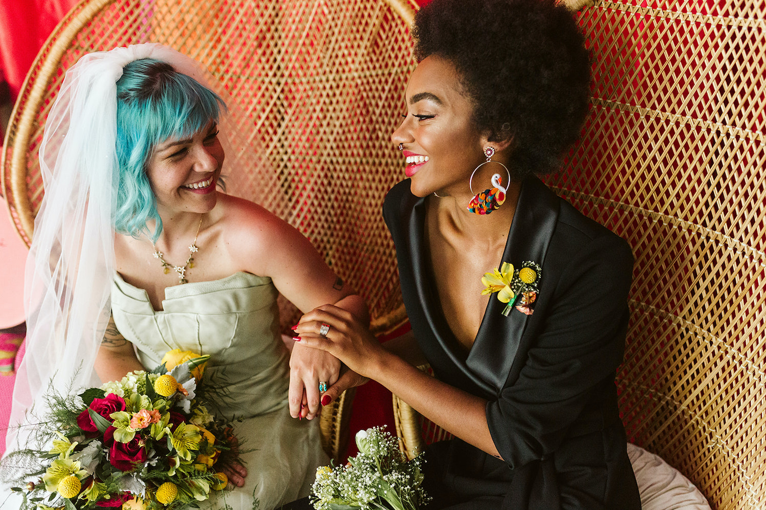 lesbian couple sits on peacock rattan chairs in front of bright wall draperies at Moxy Hotel at LGBTQ Wedding in Chattanooga