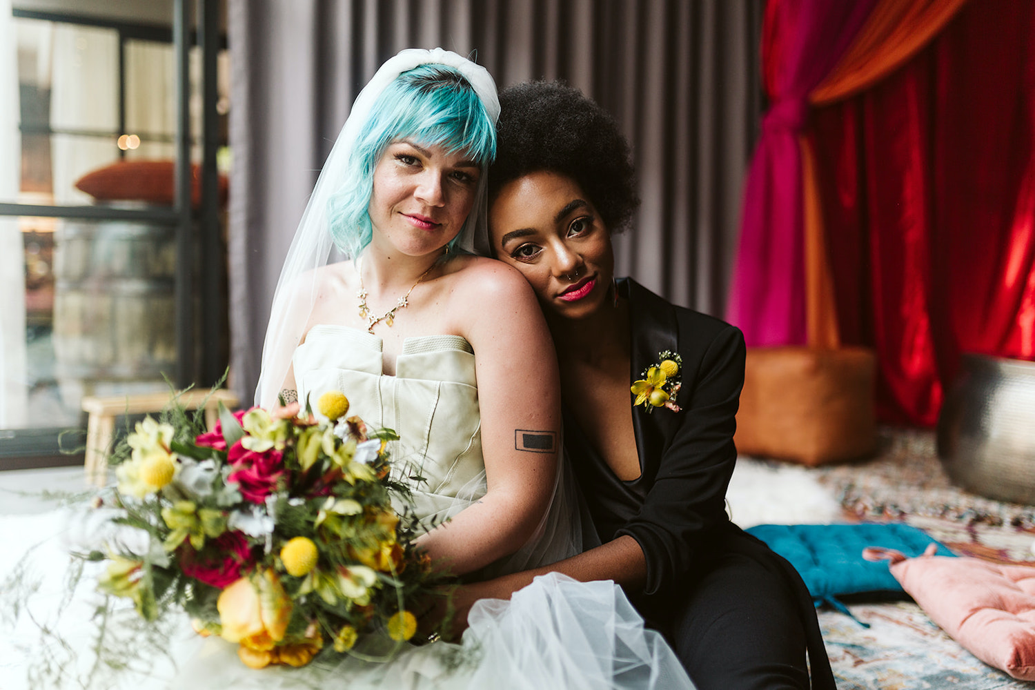 lesbian couple in strapless gown and black pantsuit cuddles on colorful pillows at Moxy Hotel at LGBTQ Wedding in Chattanooga