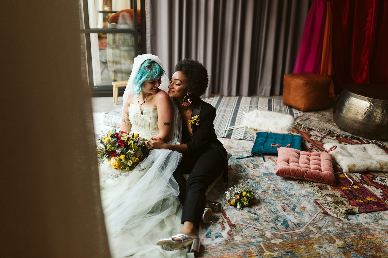 lesbian couple in strapless gown and black pantsuit cuddles on colorful pillows at Moxy Hotel at LGBTQ Wedding in Chattanooga