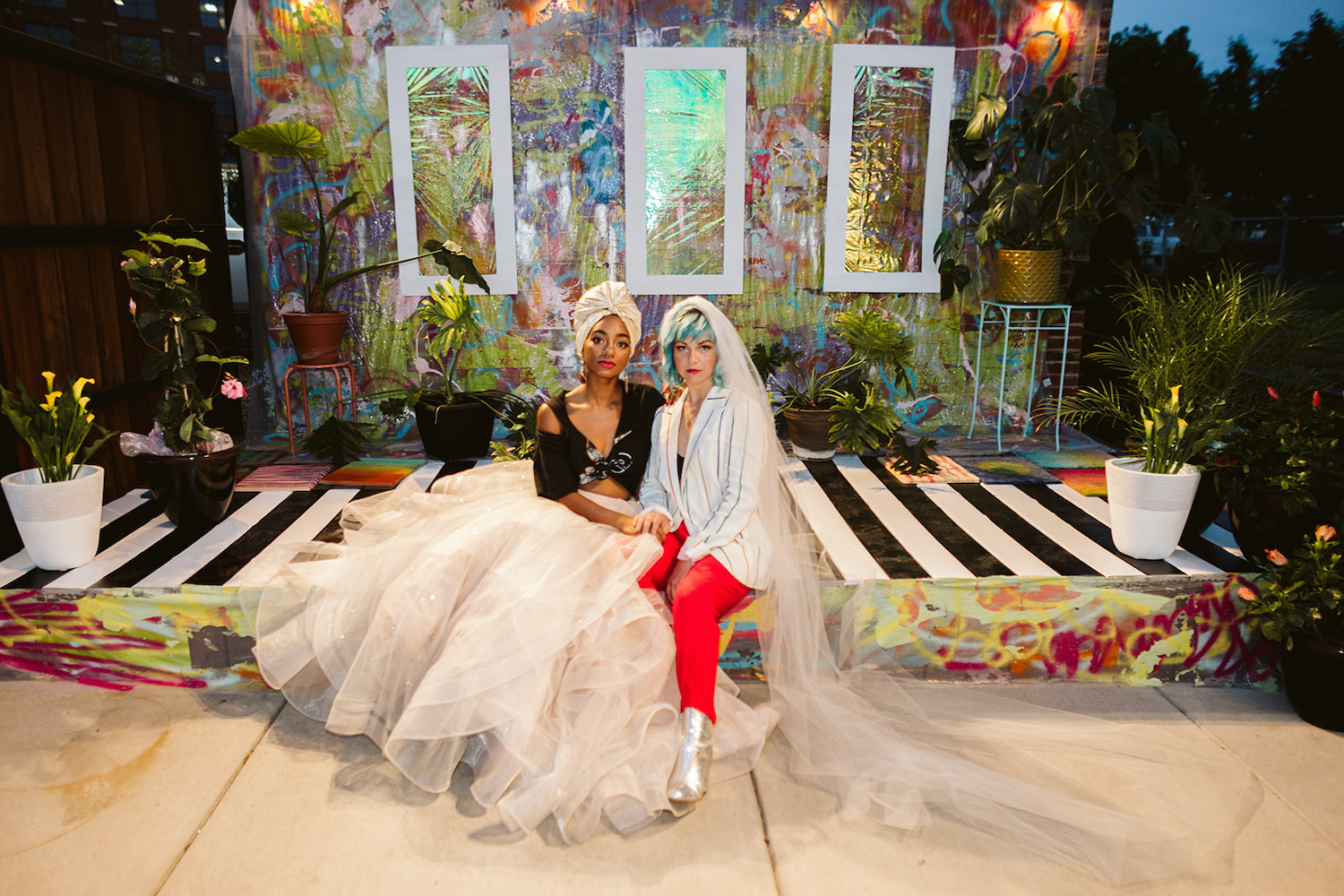 lesbian brides sit closely on an artistically painted platform with potted plants around them at LGBTQ wedding styled shoot