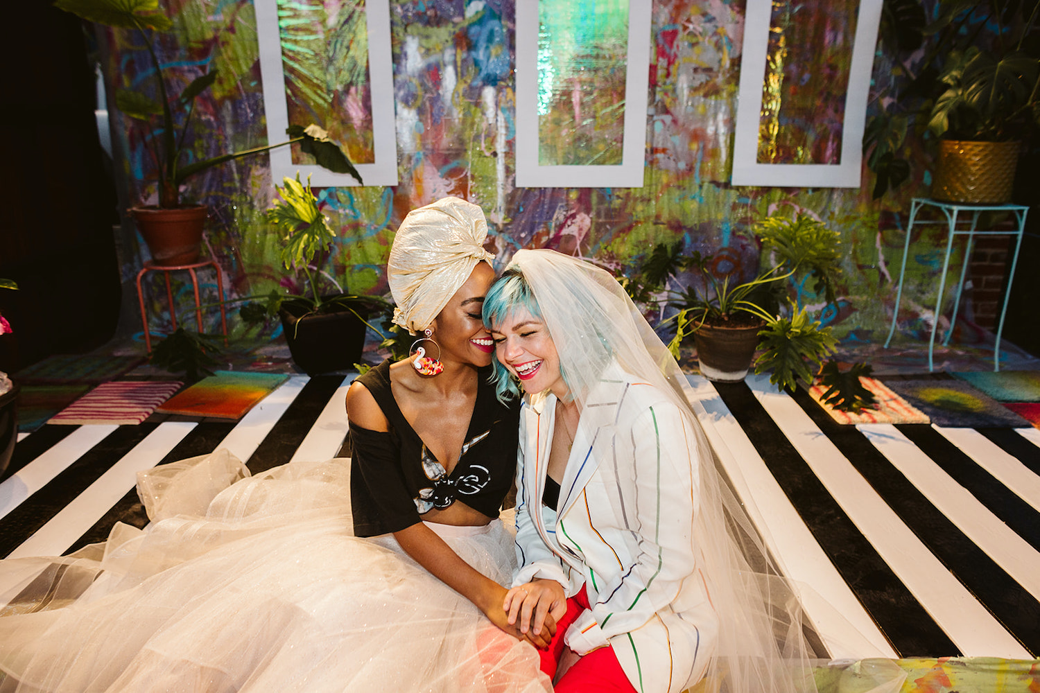 bride wearing red pants, white suit jacket and veil laughs with bride wearing full, tulle skirt, black crop top, and turban