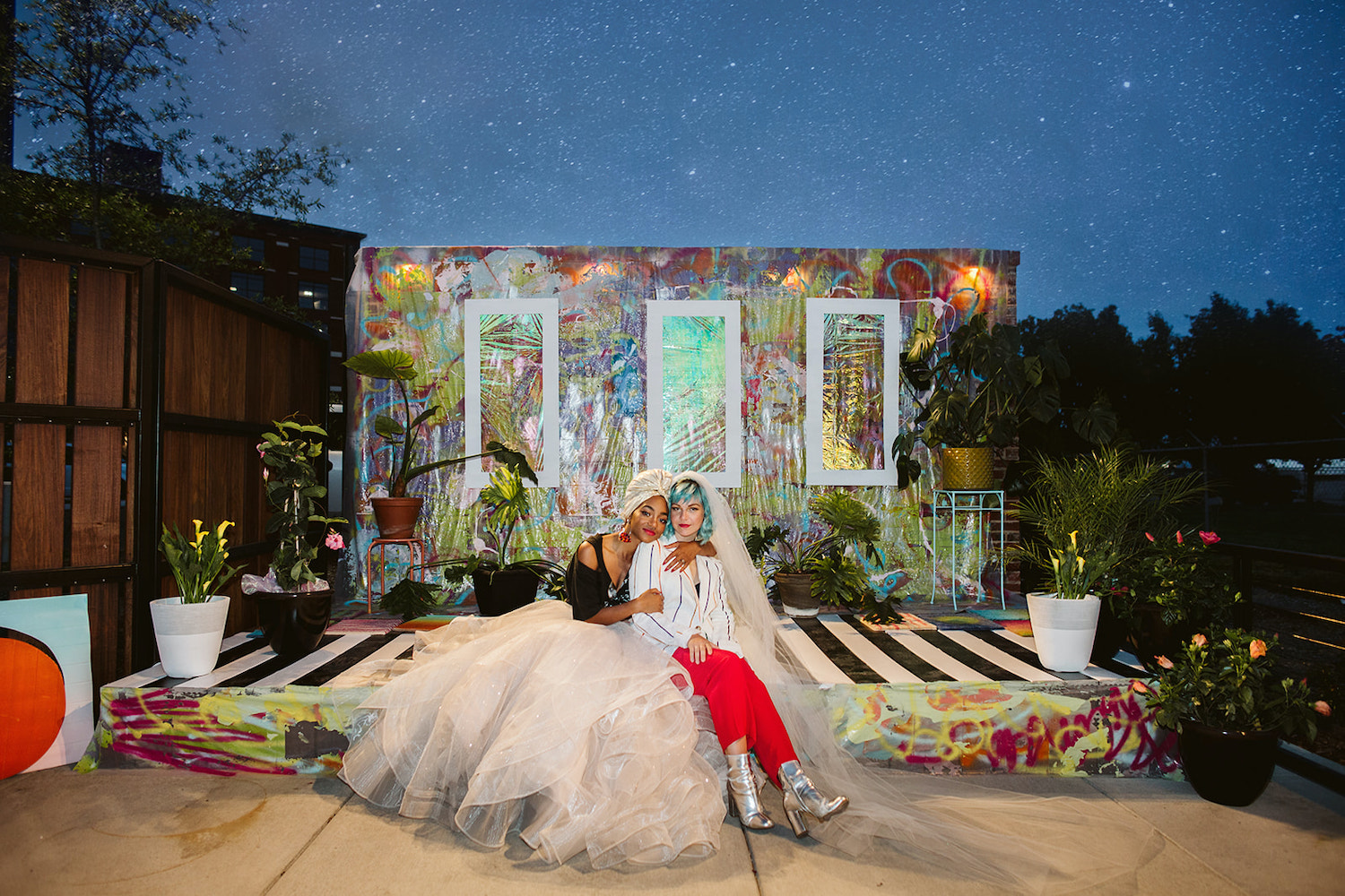 lesbian brides sit closely on an artistically painted platform with potted plants around them at LGBTQ wedding styled shoot