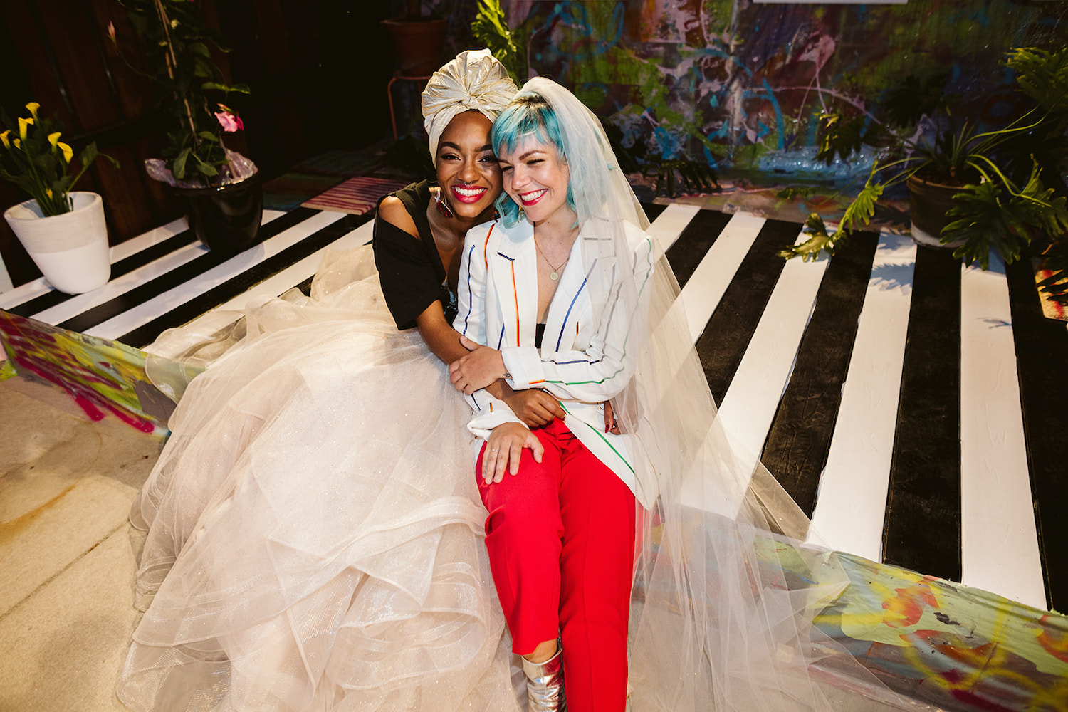 lesbian brides sit closely on an artistically painted platform with potted plants around them at LGBTQ wedding styled shoot