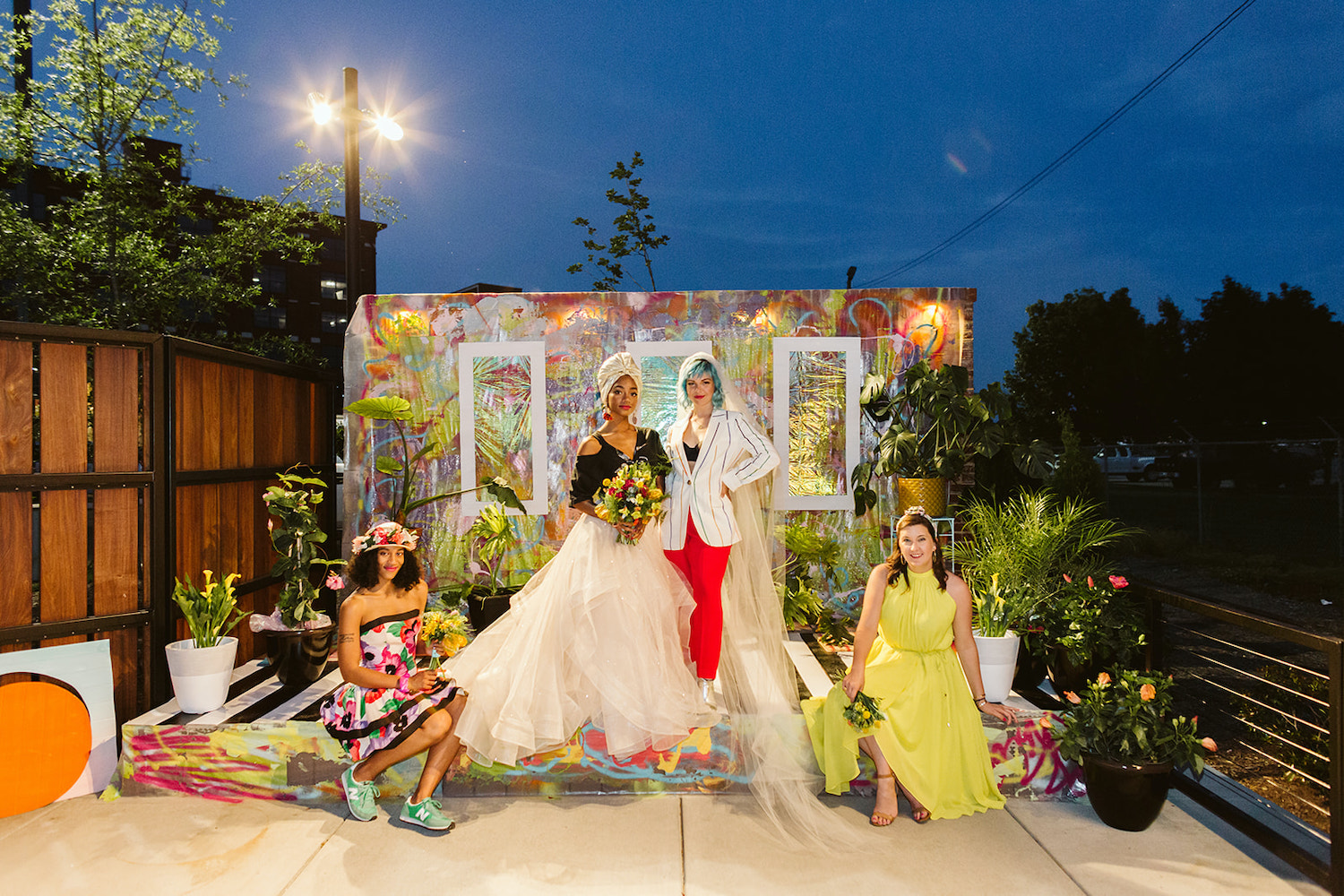 two brides and two bridesmaids wear bright, colorful wedding attire on an artistically painted platform and backdrop