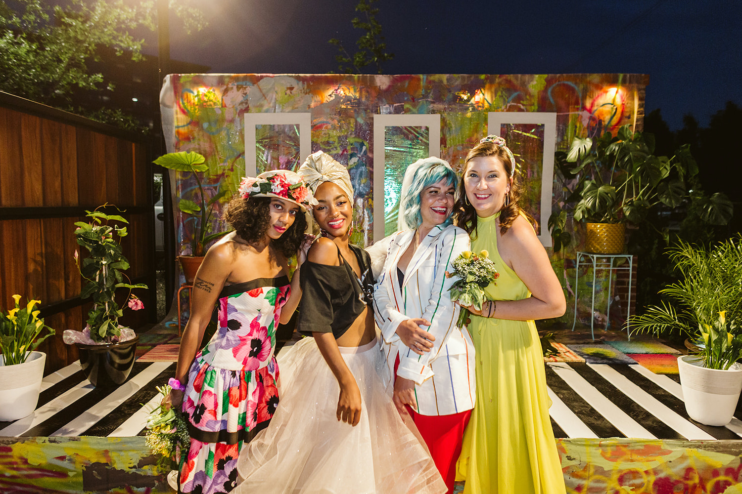 two brides and two bridesmaids wear bright, colorful wedding attire on an artistically painted platform and backdrop