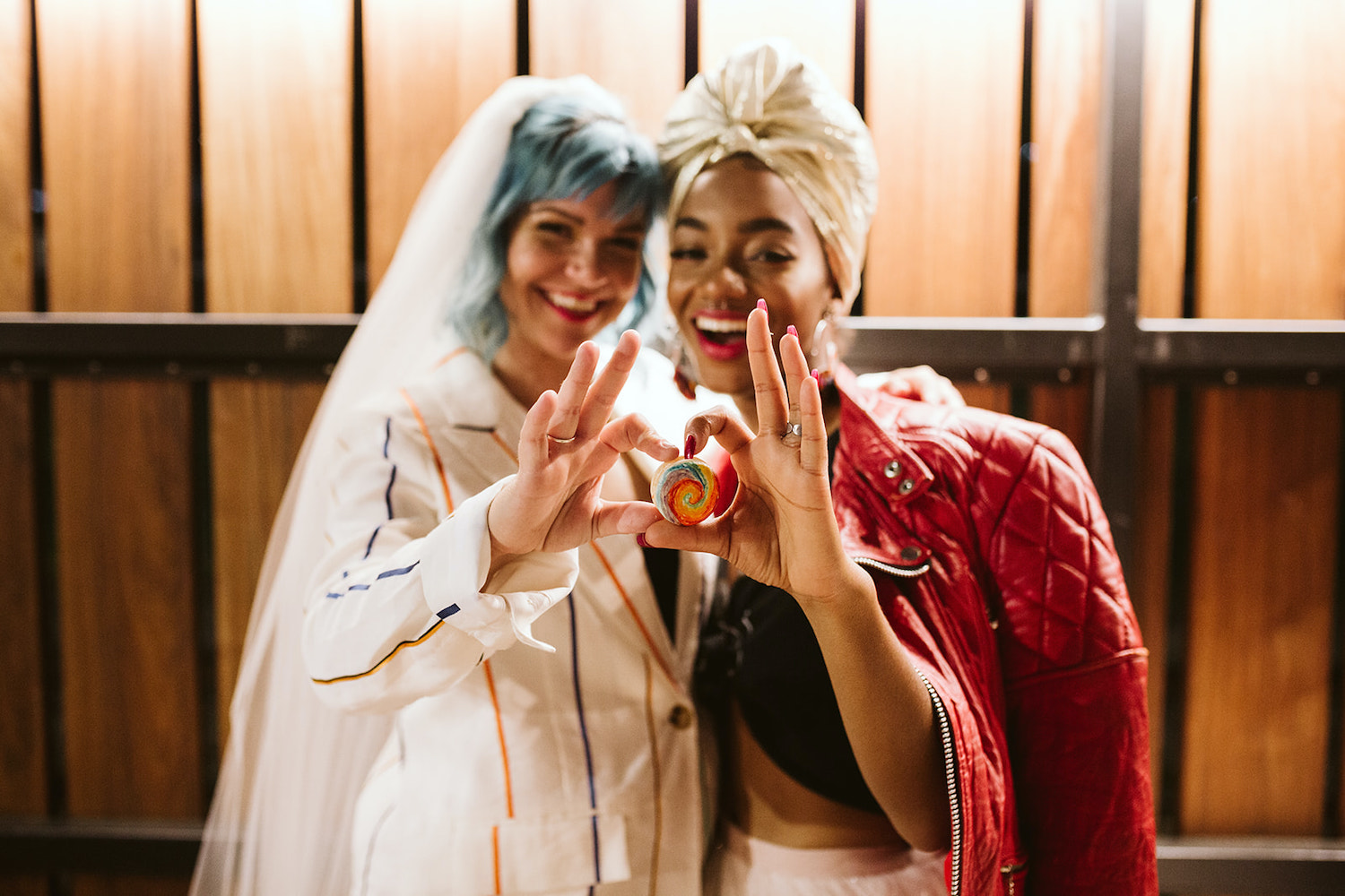 two brides stand at colorfully decorated table, holding a rainbow decorated macaroon