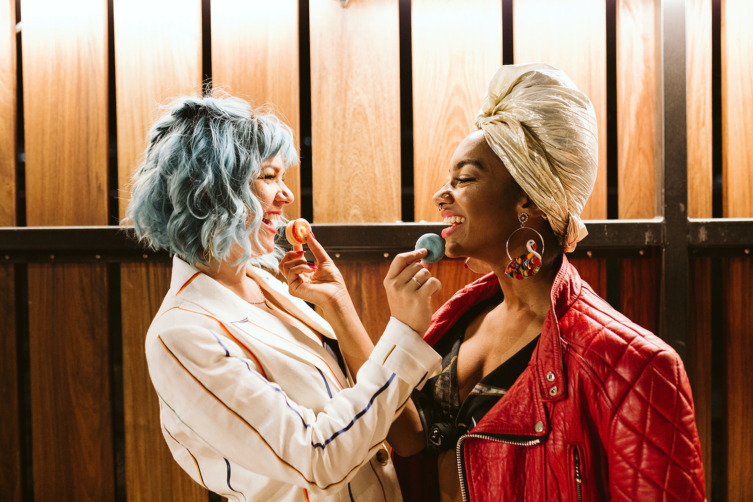 two brides stand in front of wooden fence, feeding each other macaroons