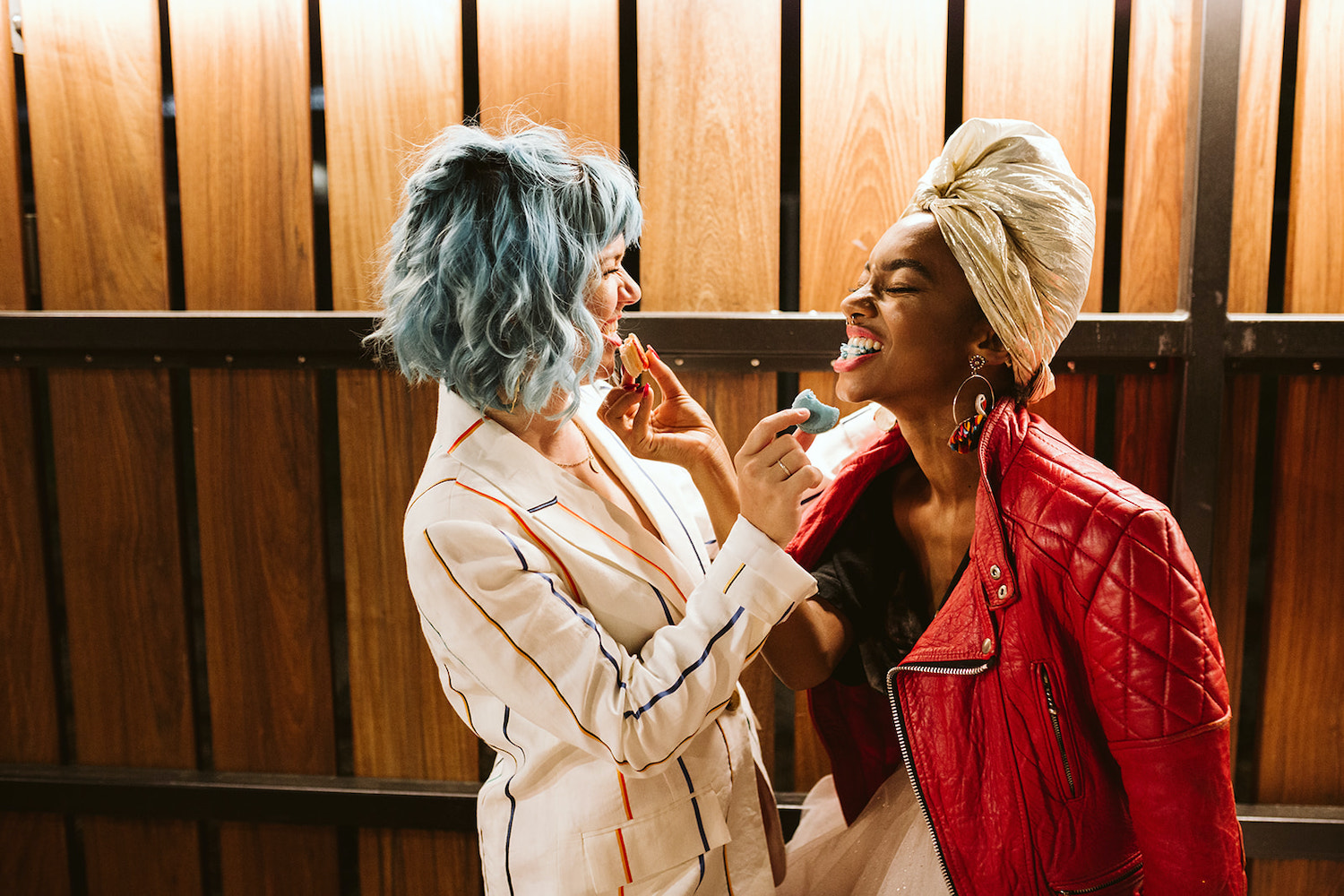 two brides stand in front of wooden fence, feeding each other macaroons