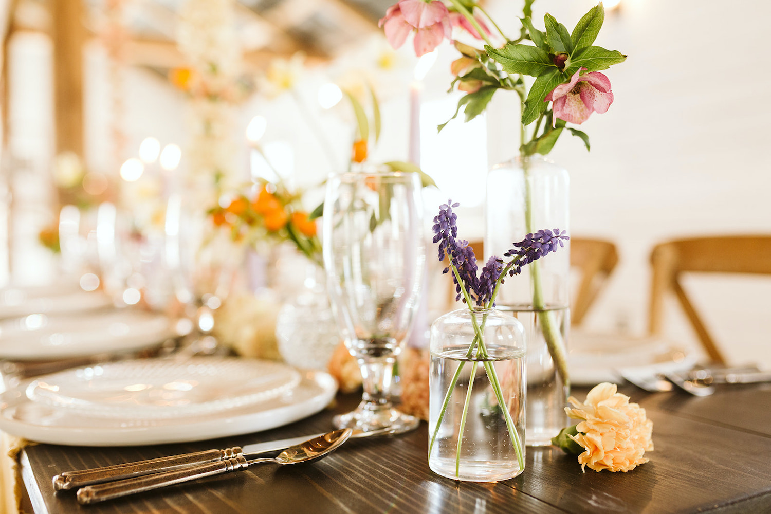 simple purple flowers in a simple glass vase on a wooden farm table next to white china and flatware