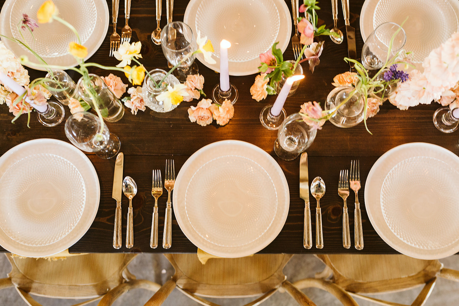 classic white place settings with flatware sits on a wooden farm table with white tapered candles and pink carnations