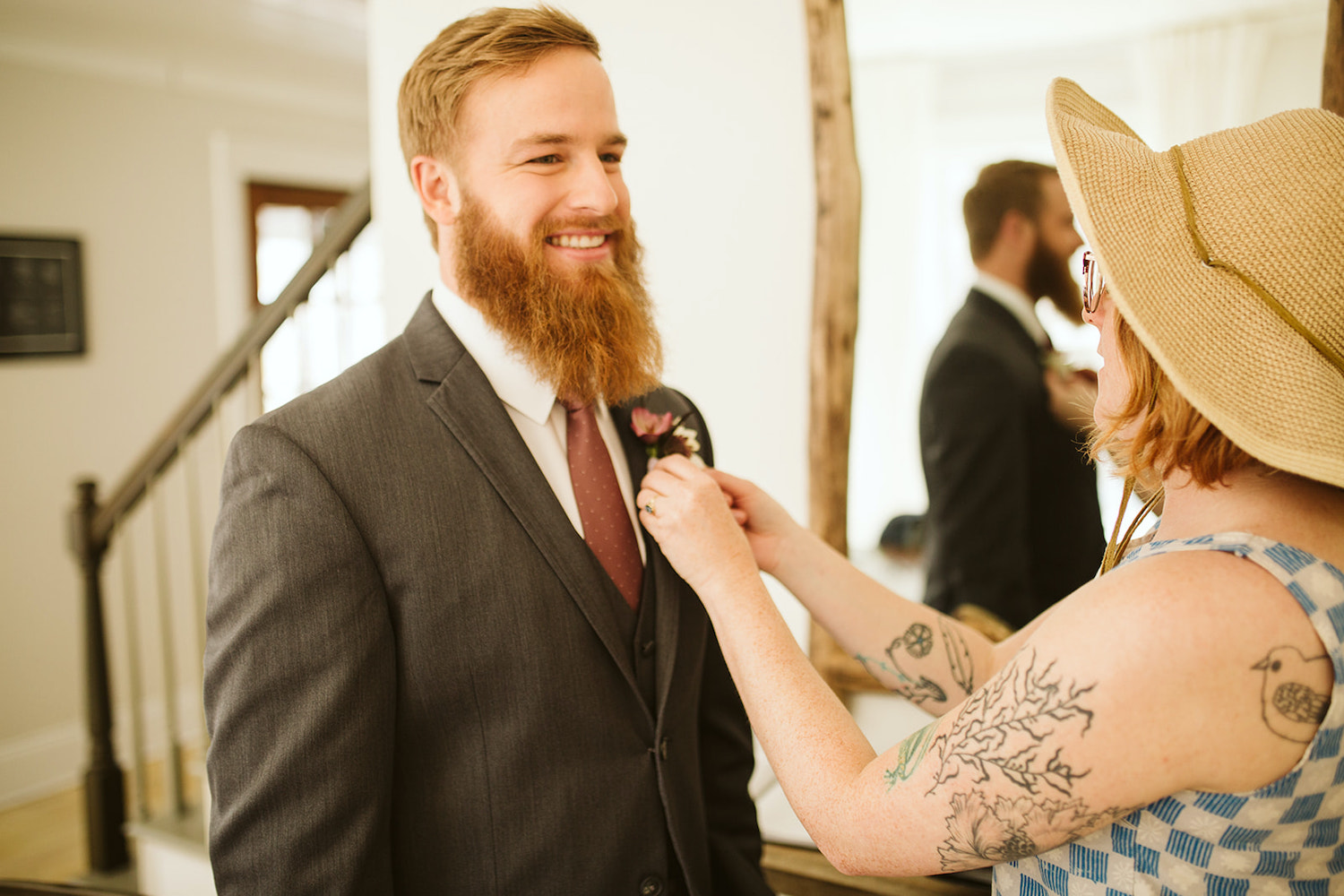 woman in blue and white checkered shirt and floppy hat pins boutonniere on groom's gray suit coat while he smiles