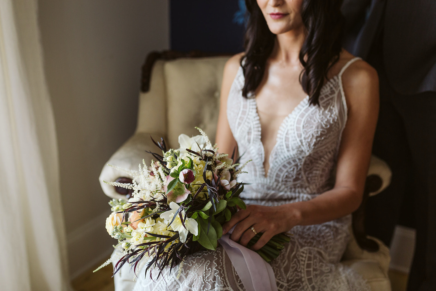 bride in embroidered wedding gown with plunging neckline holds her bouquet on a white armchair at Oakleaf Cottage wedding