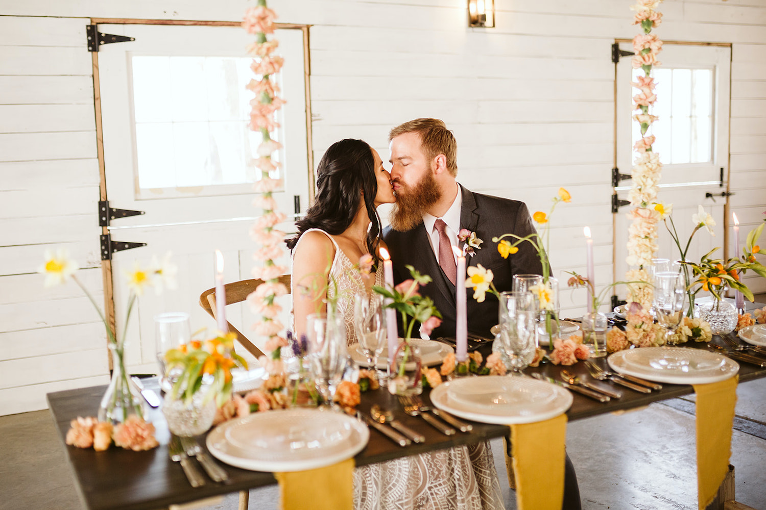 bride and groom kiss while sitting at a farm table with simple table settings and florals at Oakleaf Cottage wedding