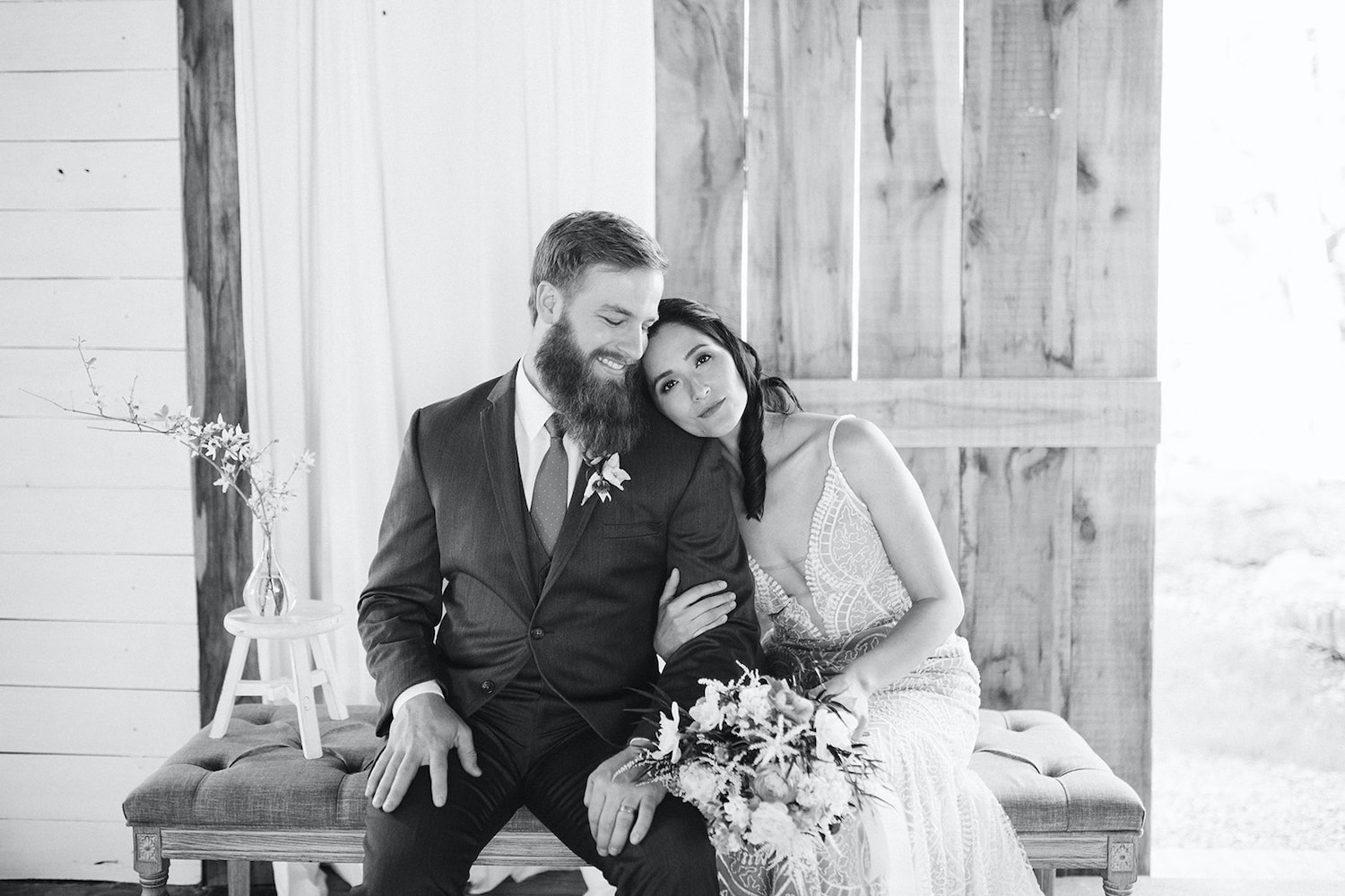 bride and groom sit on a padded bench with her hand holding his arm and her head on his shoulder at Oakleaf Cottage wedding