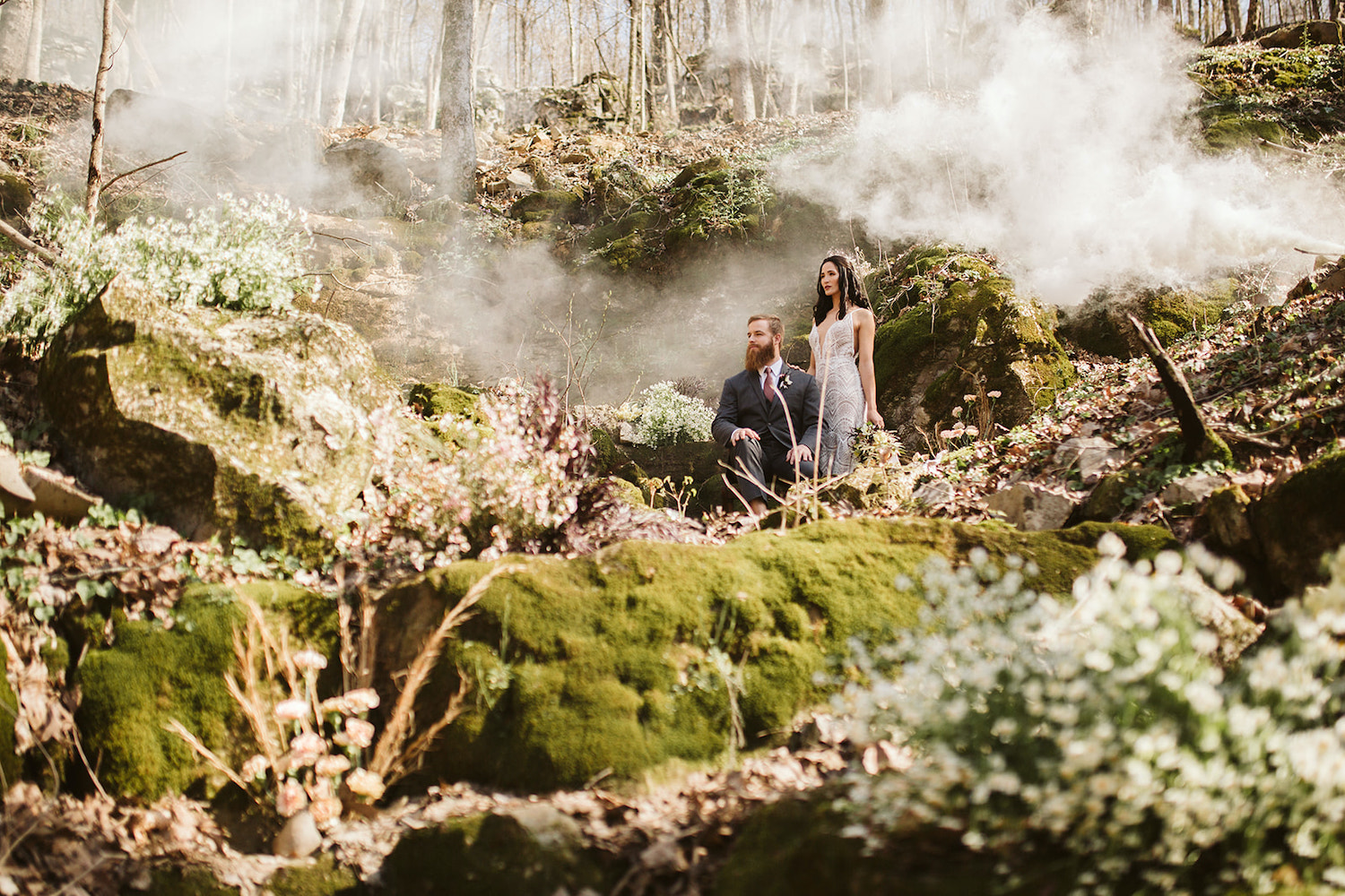 bride stands behind groom who sits on large boulder in the misty woods near their Oakleaf Cottage wedding