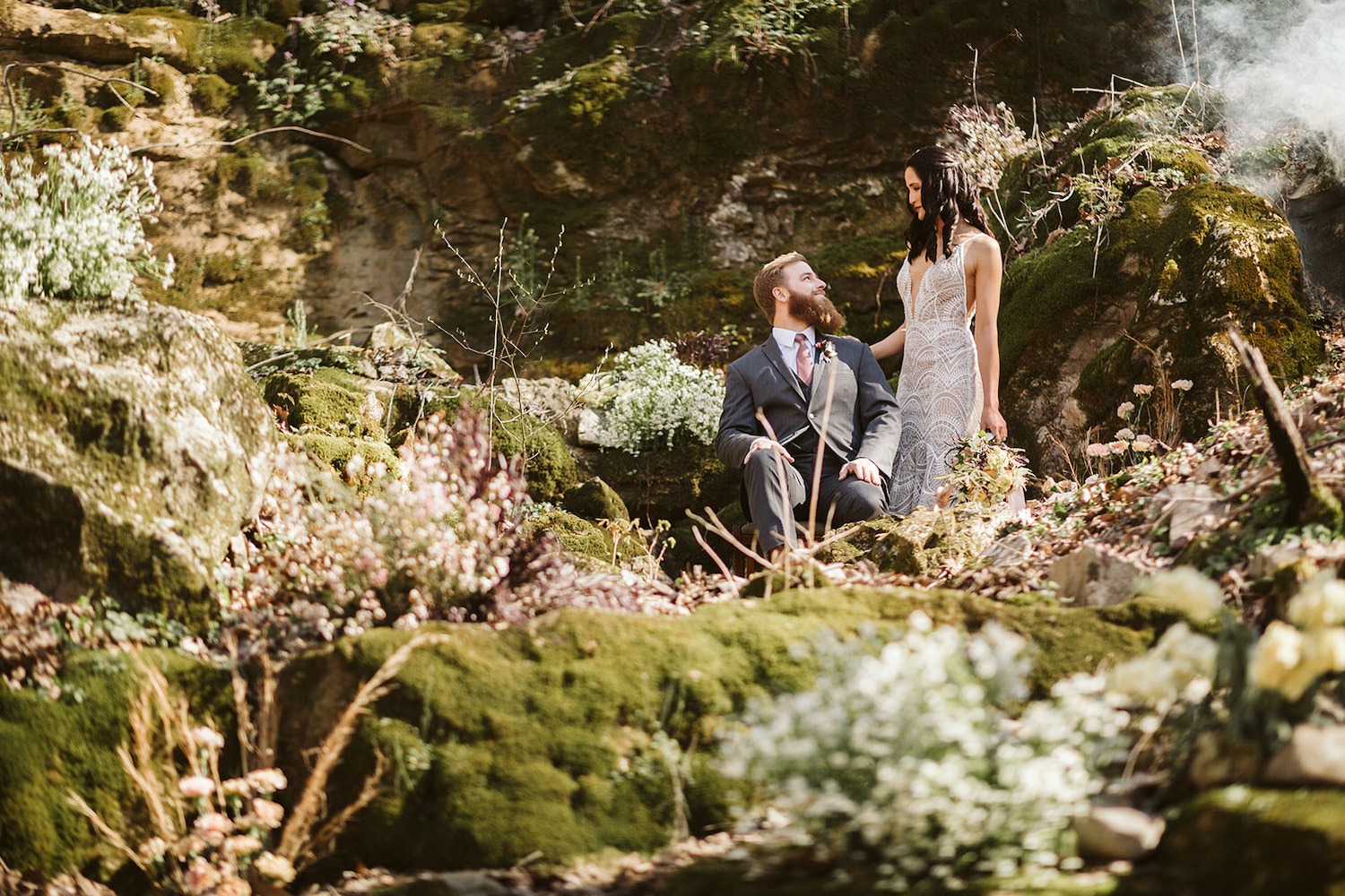 bride stands behind groom who sits on large boulder in the misty woods near their Oakleaf Cottage wedding