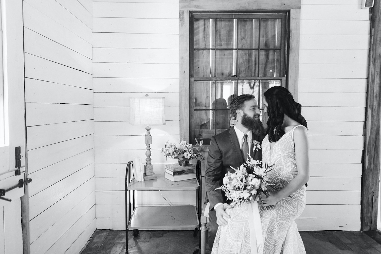 bride sits on groom's knee in front of white shiplap walls of Oakleaf Cottage wedding