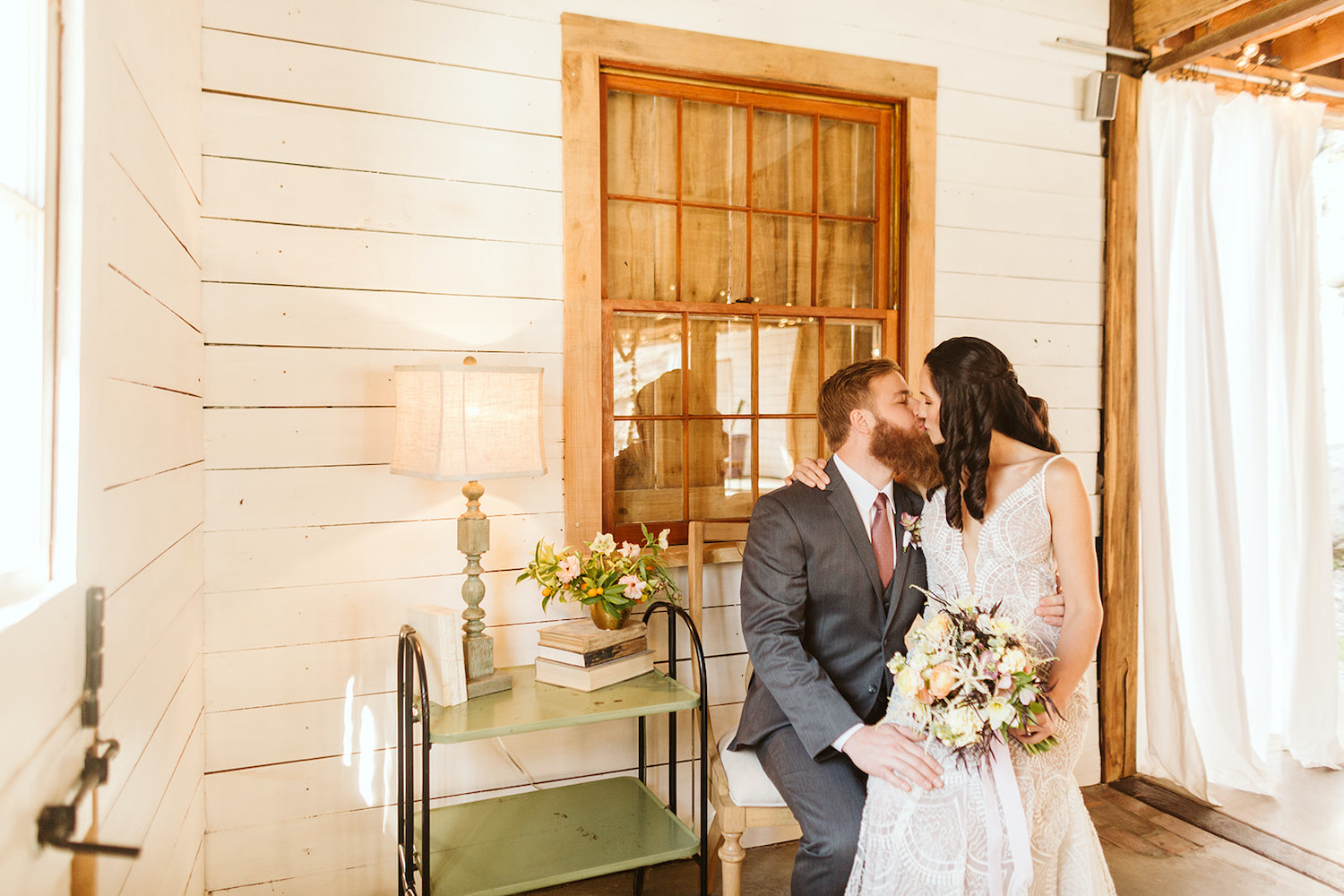 bride sits on groom's knee as they kiss in front of white shiplap walls of Oakleaf Cottage wedding