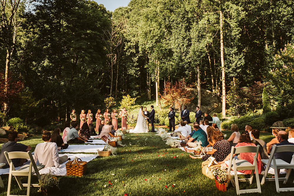 Wedding guests sit on blankets in a meadow as the bride and groom exchange vows.