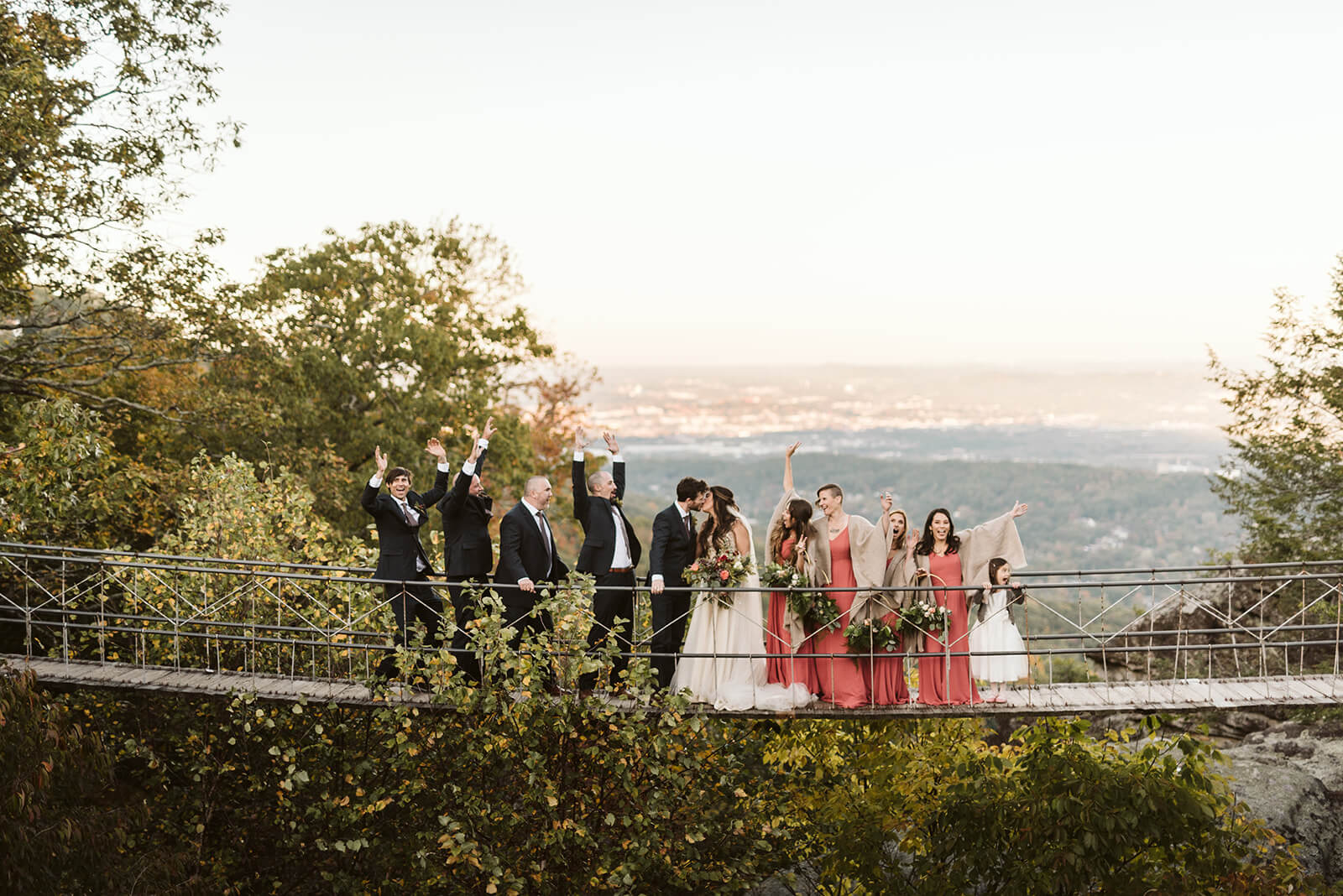 A bridal party celebrates as the bride and groom kiss on a suspension bridge.