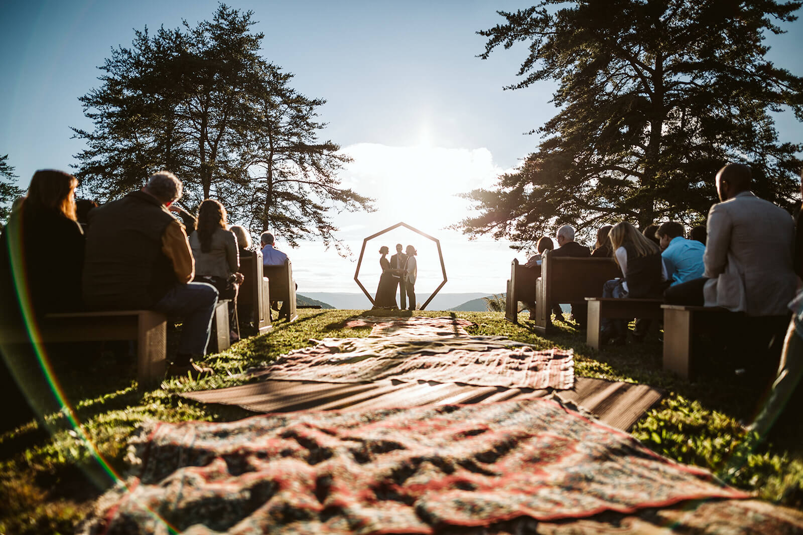 A bride and groom exchange vows under a geometric arbor at sunset. The aisle is lined with antique rugs.