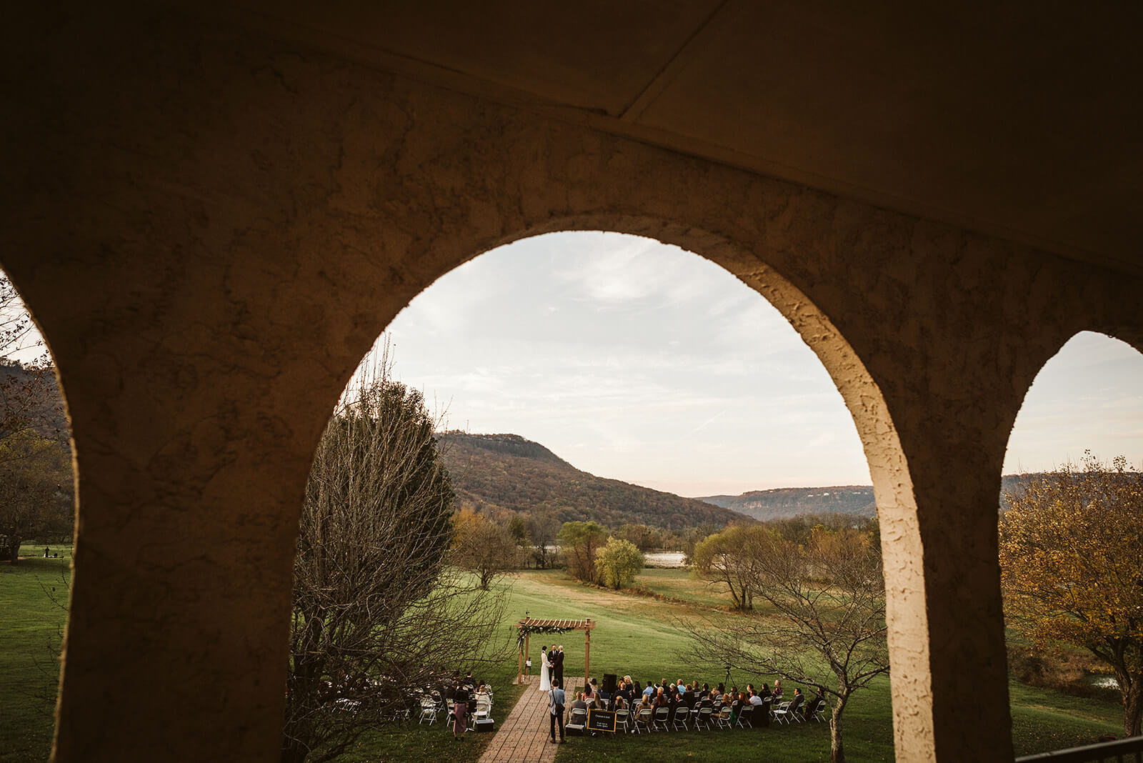 A shot through a Spaniish-style arch of a bride and groom exchanging vows in a field next to a river.