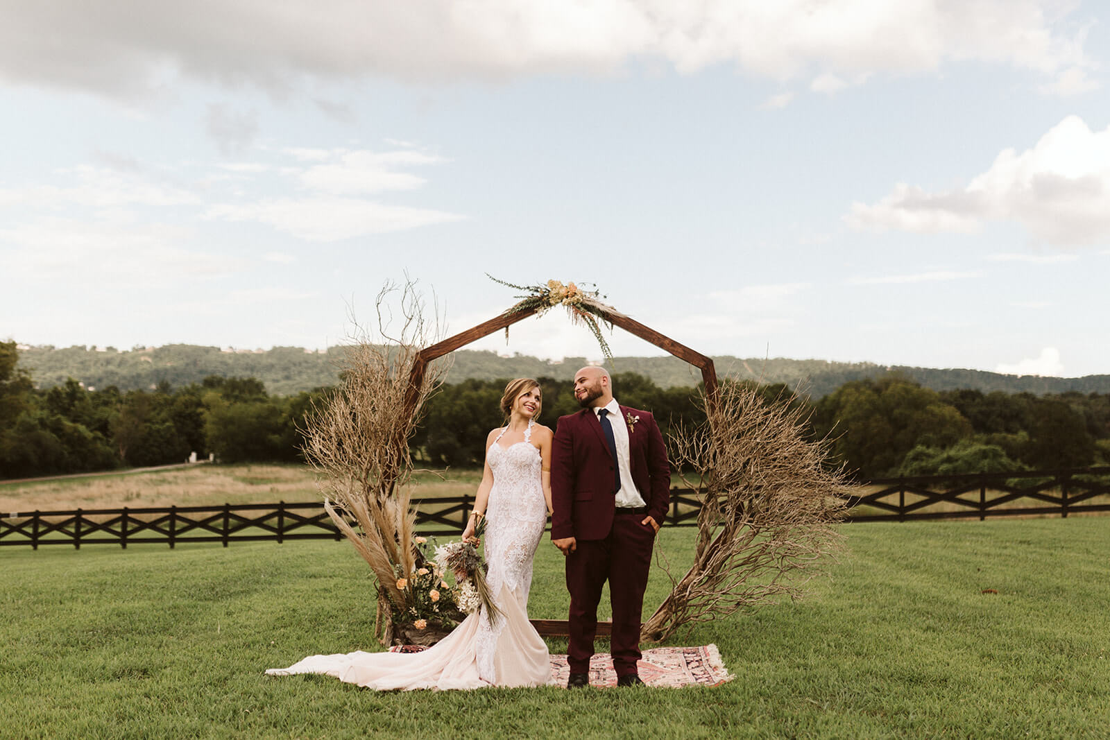 A bridge and groom stand together under an octagon arbor in a field.