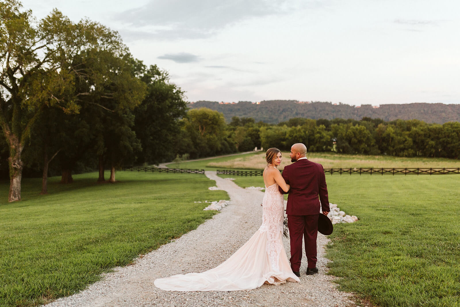 A bride and groom walk arm-in-arm down a gravel road through an open field.