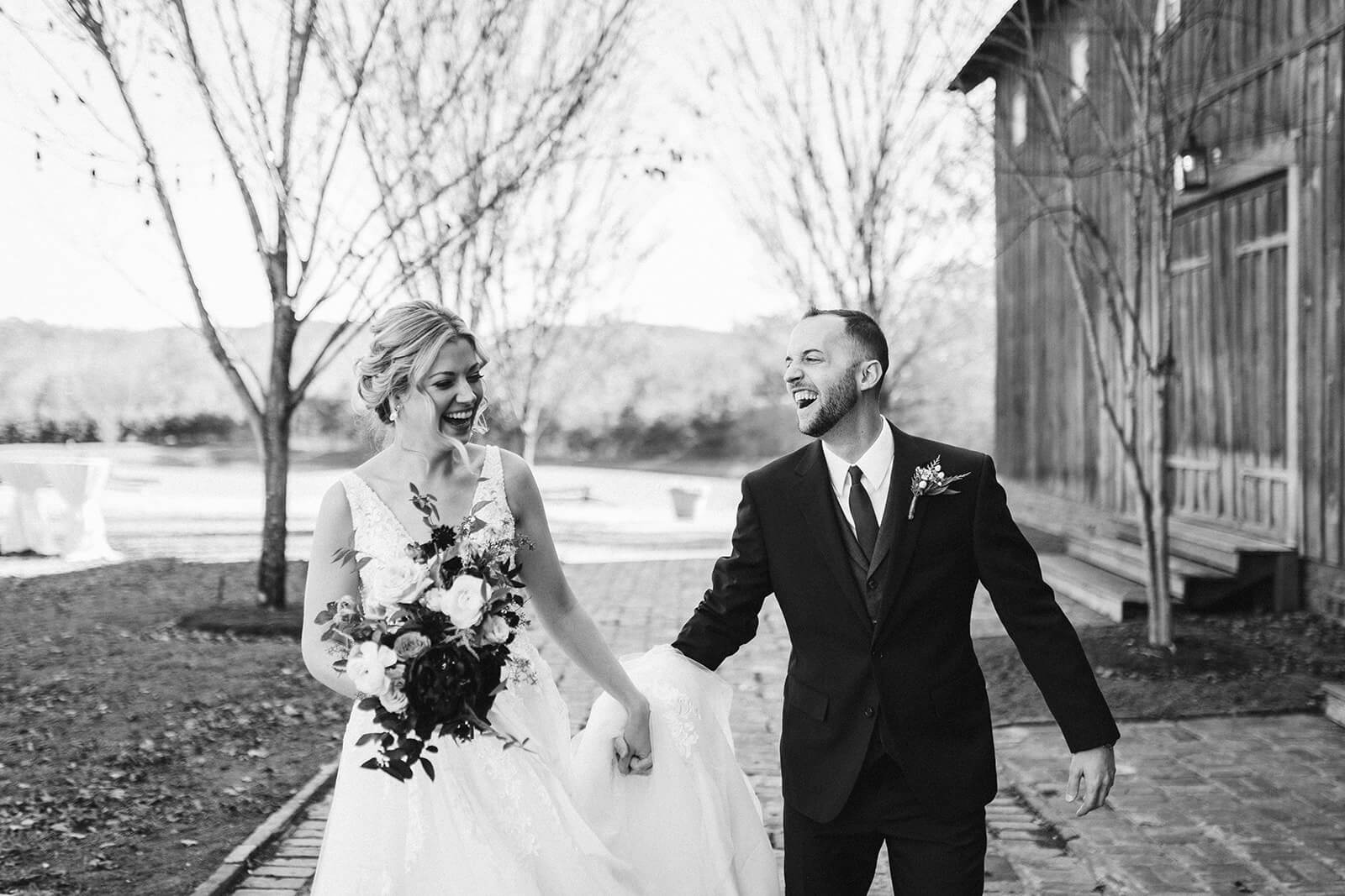 A black and white photo of a bride and groom walking hand-in-hand down a brick path by a barn.