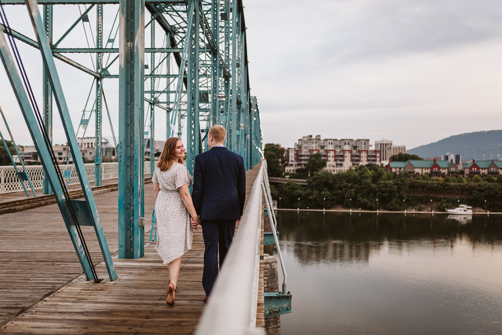 A couple crosses the Walking Bridge in Chattanooga, TN, hand-in-hand. The woman looks back over her should and smiles at the camera.