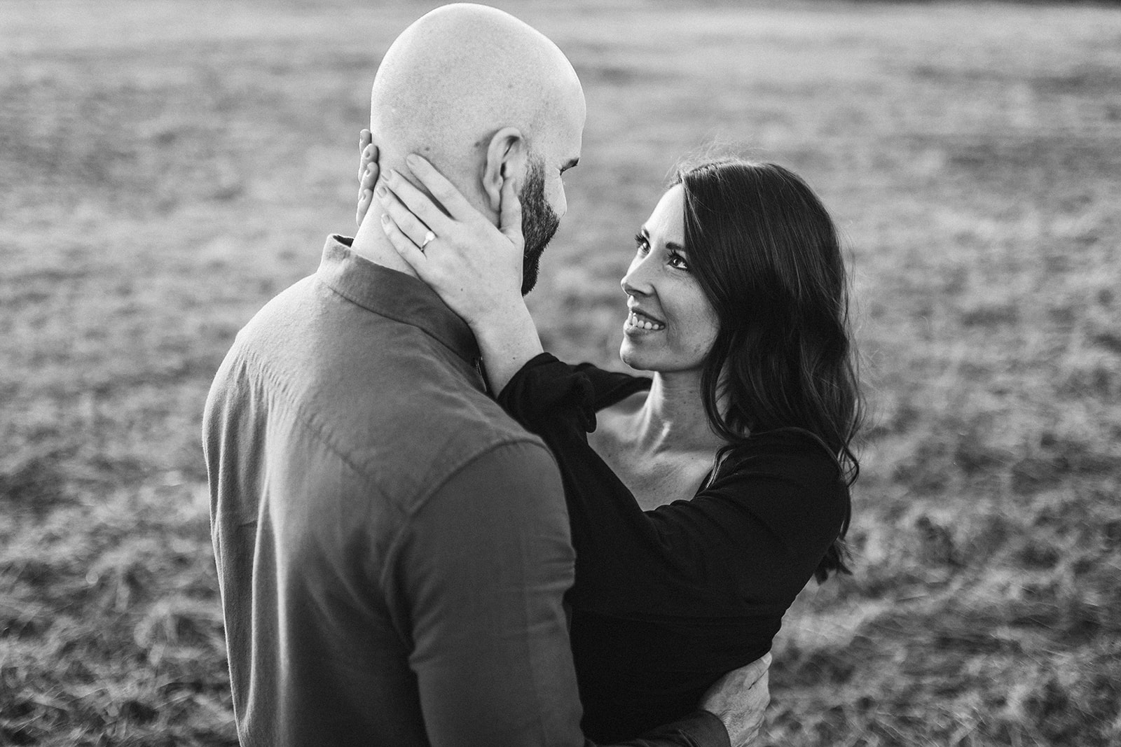 A black and white photo of a woman holding her fiance's face and looking up into his eyes.