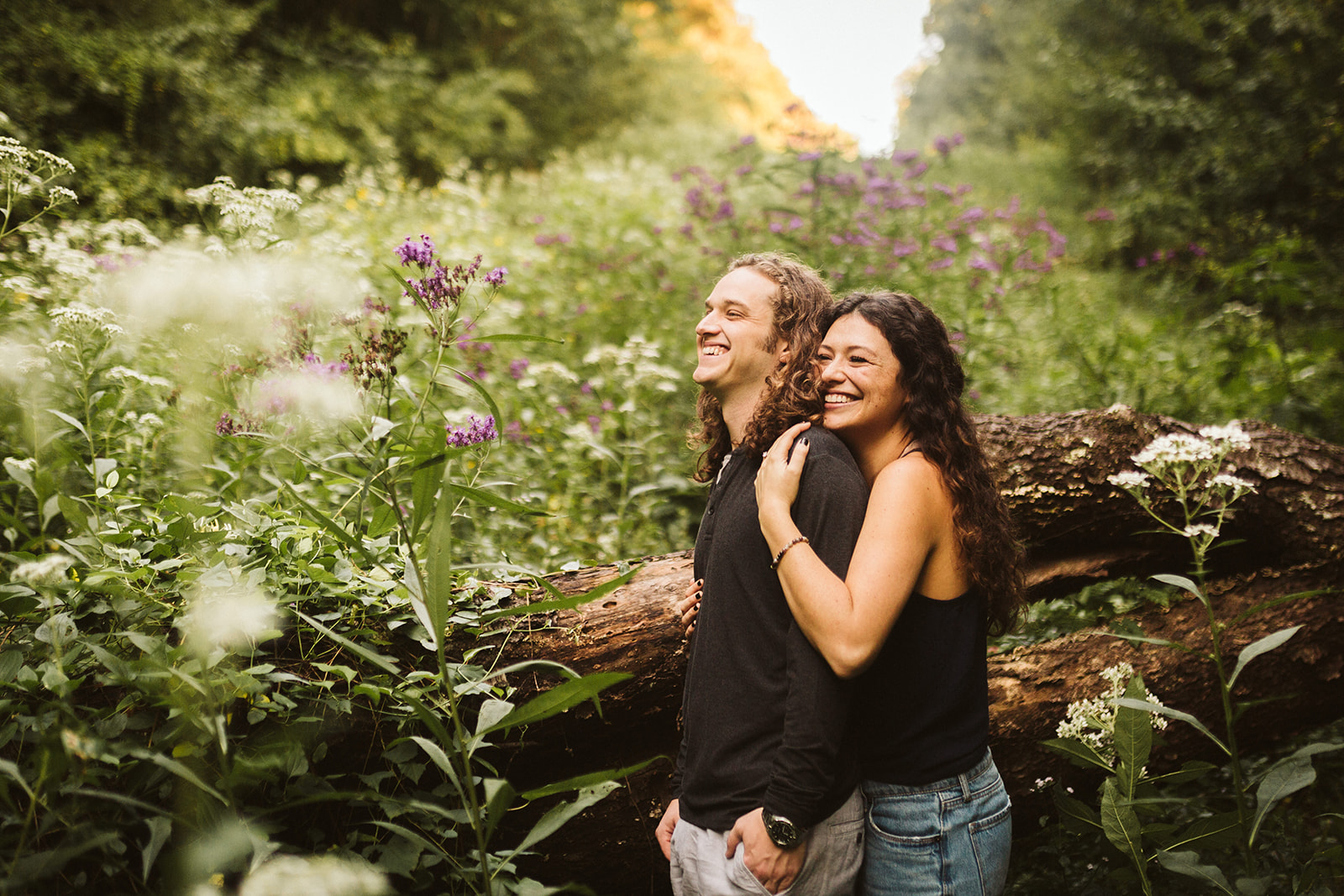 A woman hugs her fiance from behind in a field of wildflowers while they stand for an engagement photo pose.