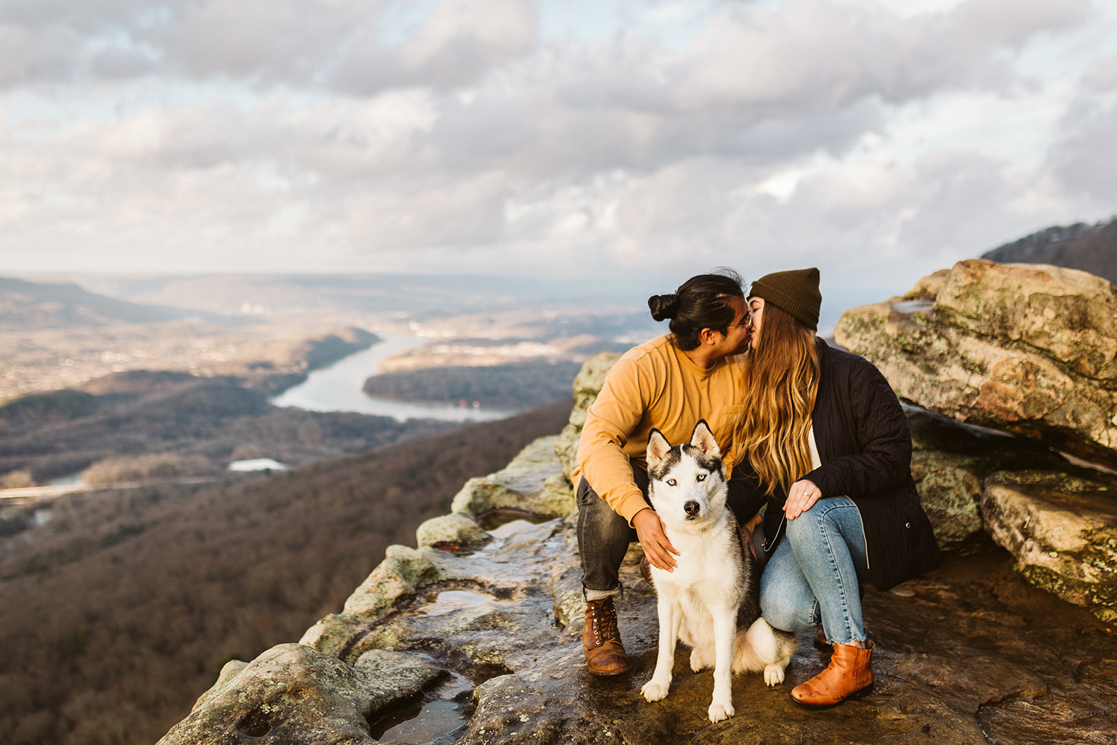 An engaged couple kisses while holding their husky on top of a mountain.