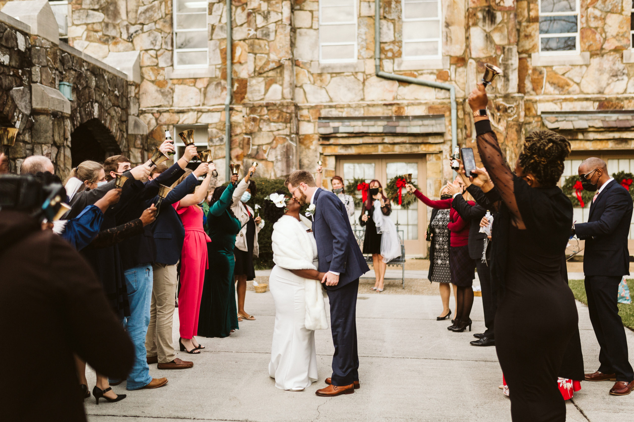 A bride and groom kiss in front of a stone building while their guests stand around them ringing bells.