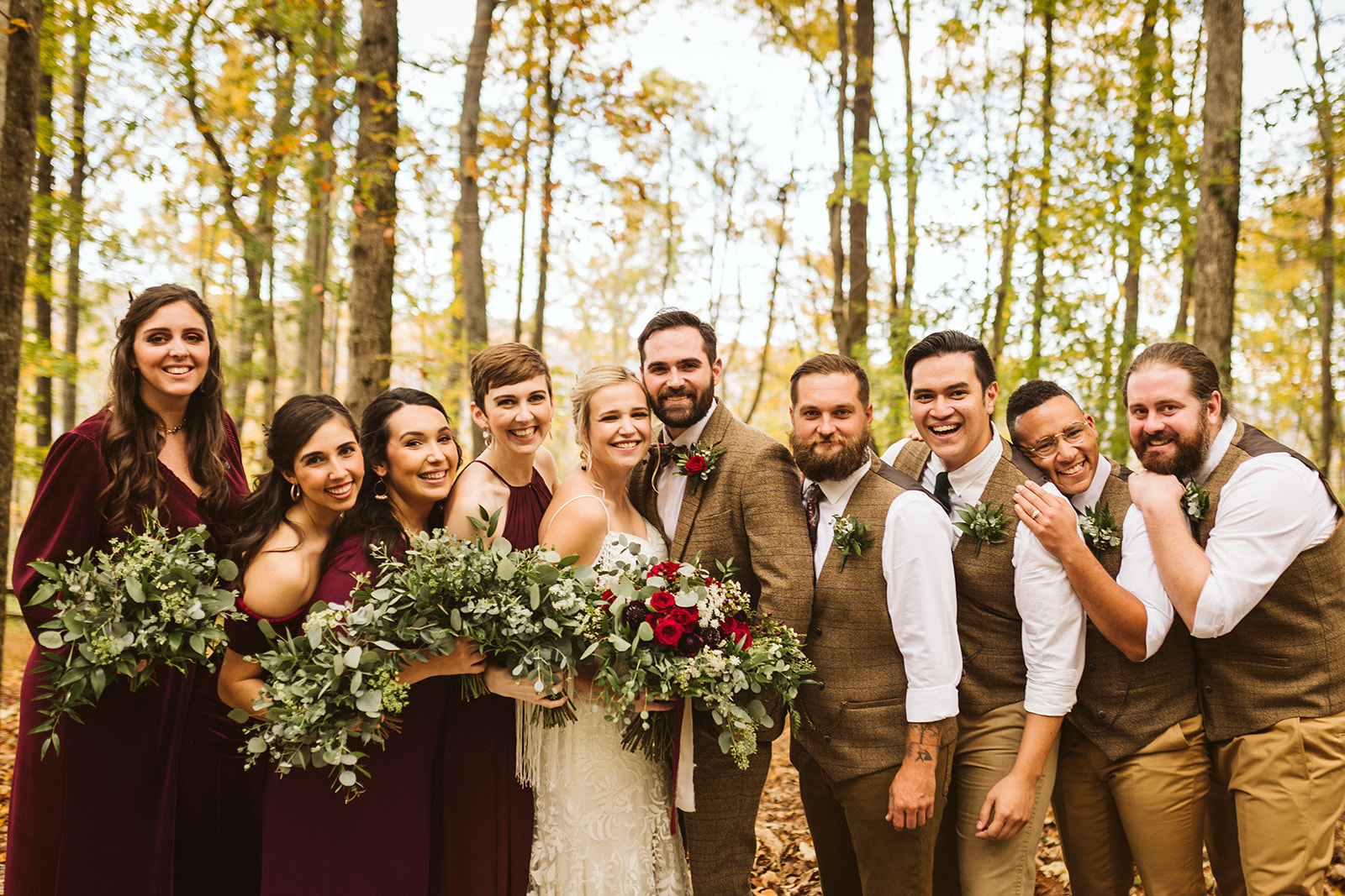 A bride and groom pose with their bridal party in a forest in fall.
