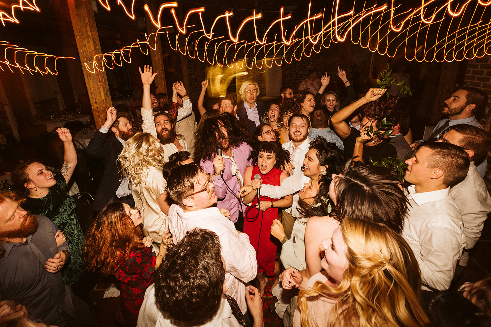 Wedding guests dance and sing into microphones under blurry lights.