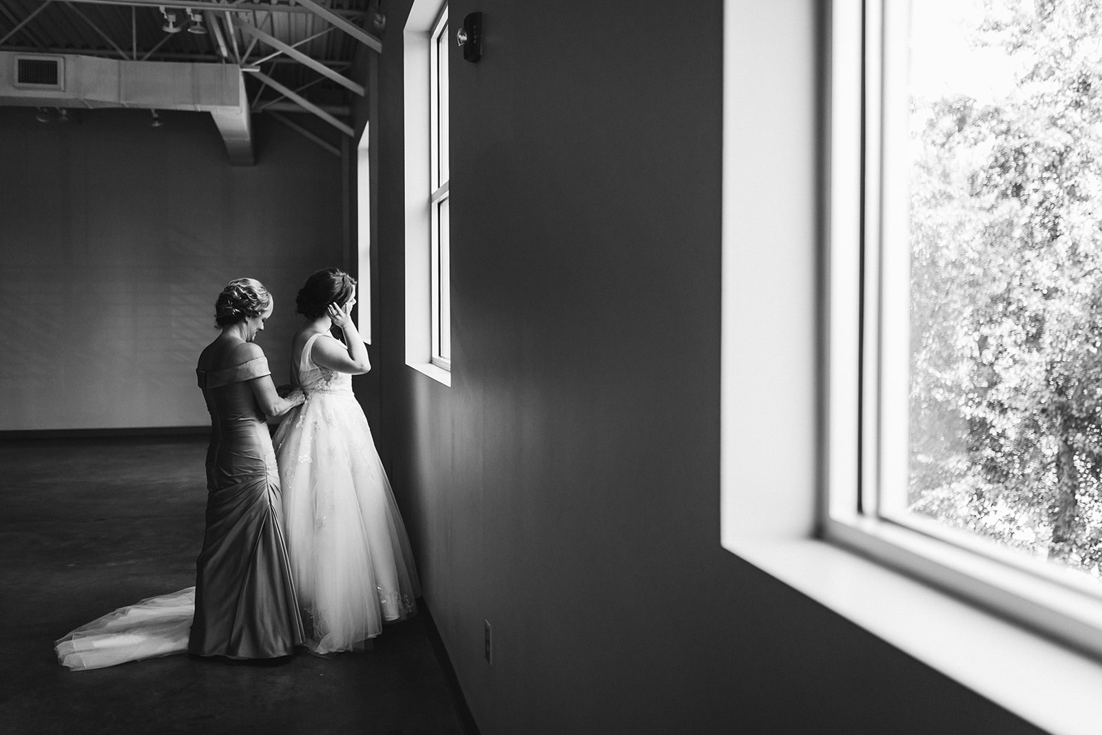 A black and white photo of a bride's mother buttoning her dress in front of a bright window.